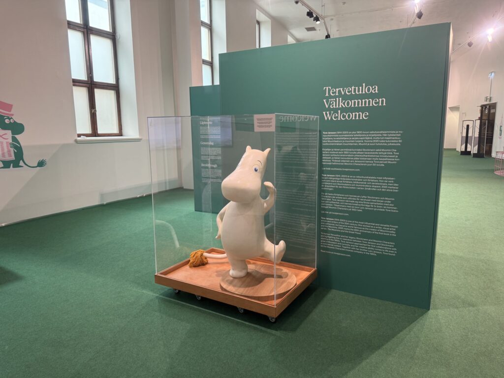 A large statue of Moomintroll inside a glass display case at a Moomin exhibition. The statue is on a green carpeted floor, with a large green wall in the background that has 'Welcome' written on it in several languages.