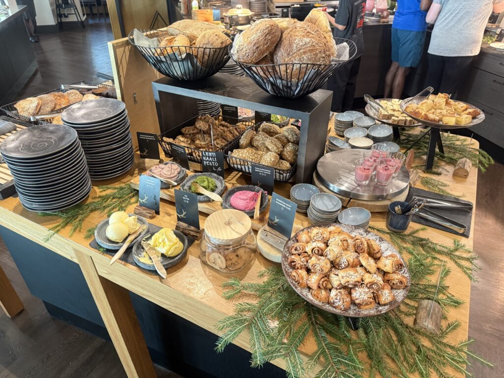 A hotel breakfast buffet. Various types of bread in baskets, pastries, colorful dips, and butter are beautifully arranged on a wooden table.