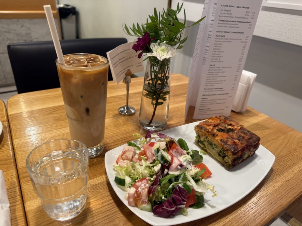 A plate of quiche and green salad, a glass of iced caffè latte, a glass of water, and a vase with flowers on a wooden table.