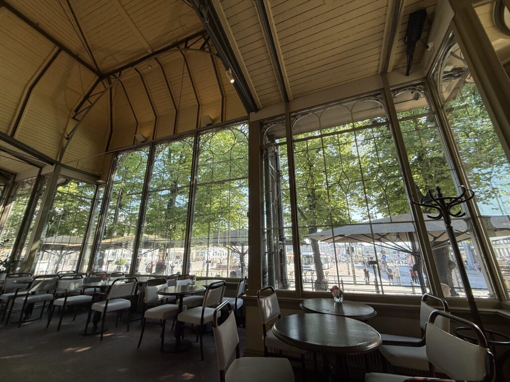 The interior of a classic cafe with a high, arched ceiling and large windows looking out onto lush green trees. Tables and chairs are arranged inside.