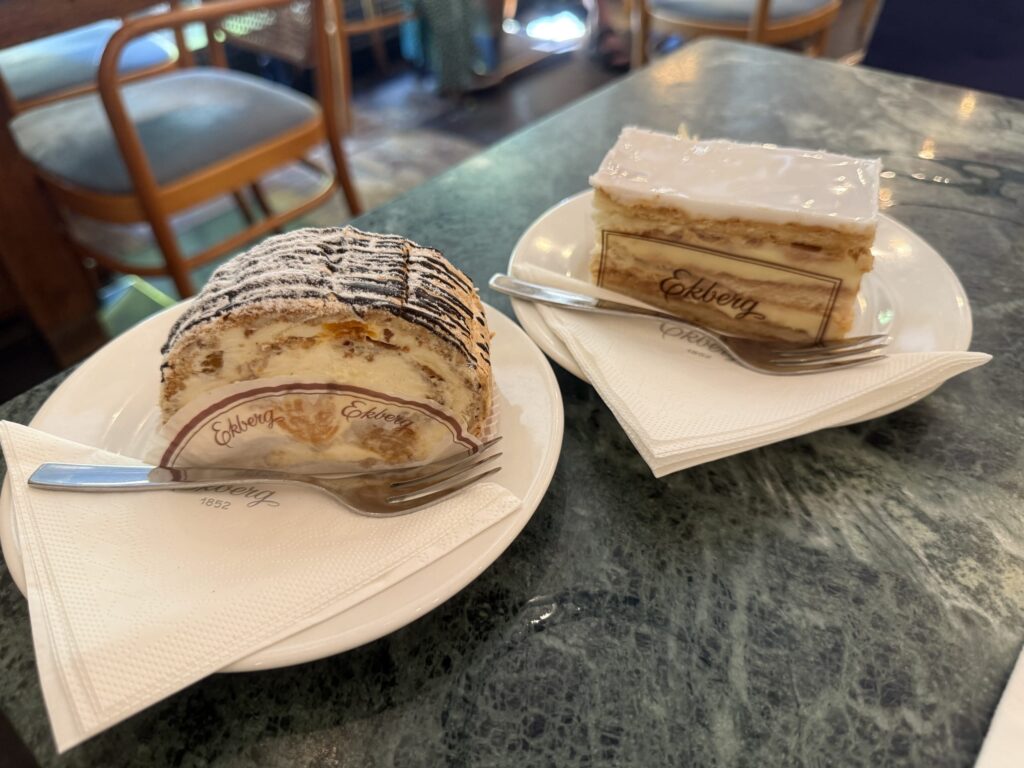 Two types of cakes from the historic Ekberg café in Helsinki on white plates, placed on a green marble table. In the foreground is a cream-filled roll cake, and in the background is an Alexander Torte with white icing.