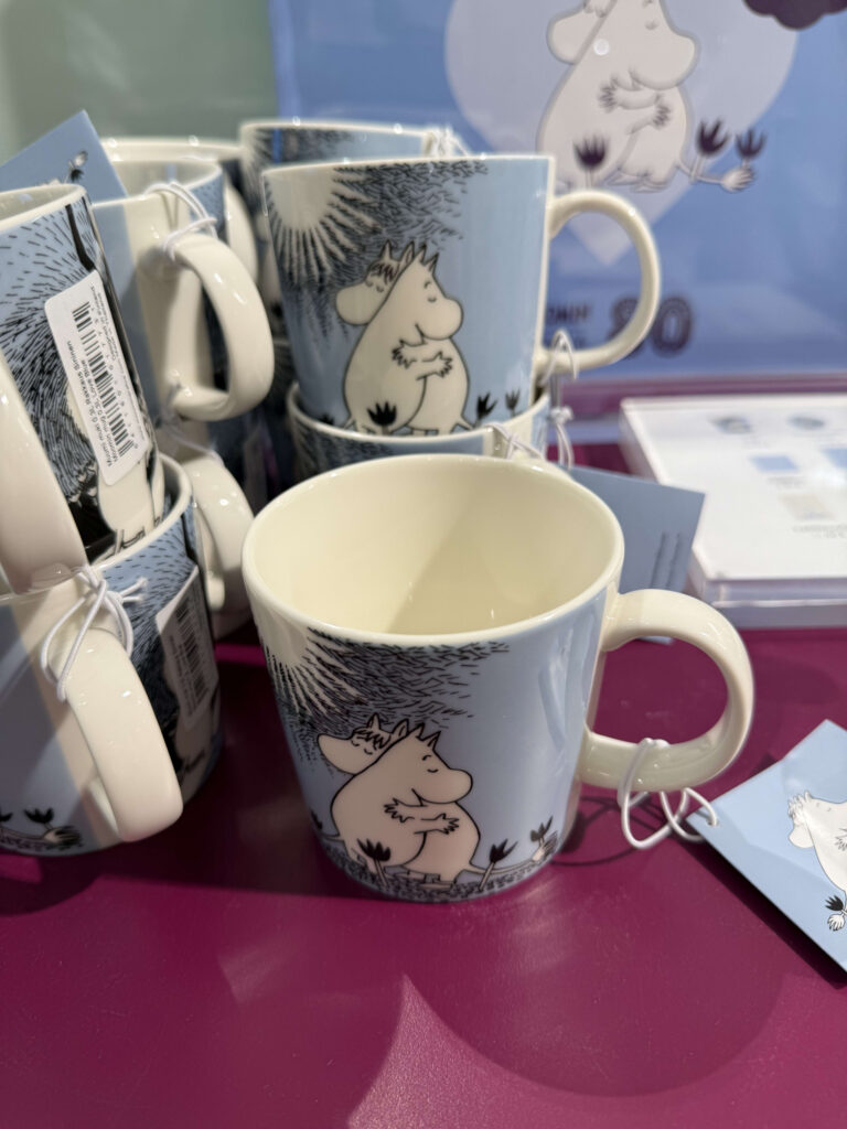 A collection of light blue Moomin mugs with an illustration of two characters hugging, displayed on a bright magenta table. The mug in the foreground is in sharp focus.