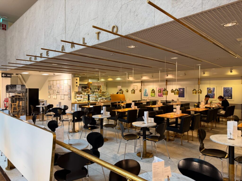 The spacious interior of Cafe Aalto, located inside the Academic Bookstore. The cafe's name is displayed in golden letters on a marble wall, and numerous brass pendant lights hang from the ceiling. Black chairs and tables are arranged, with a few customers relaxing.