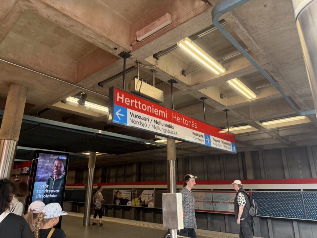 The platform of Herttoniemi metro station in Helsinki, Finland. A red and white sign with the station's name hangs from the concrete ceiling, as several passengers wait for a train.