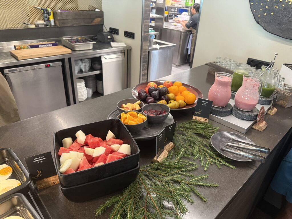 A fruit corner at a hotel breakfast buffet. Cut watermelon, oranges, apples, and other fruits are lined up on the counter, along with pink and green smoothies.