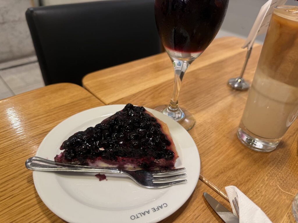 木製のテーブルの上にあるカフェ・アアルトの白い皿に乗せられたビルベリータルトのスライス。隣には赤いジュースとアイスラテが置かれている。