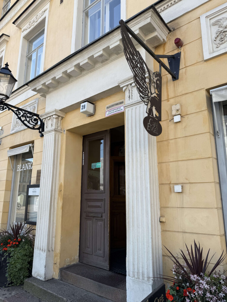 The entrance to Café Engel, with its yellow facade, white columns, and a prominent black sign shaped like an angel's wing above the door.