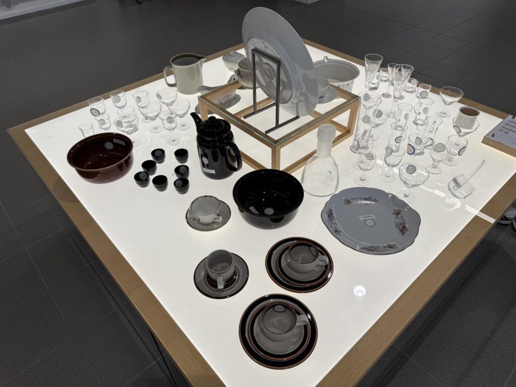 A collection of vintage tableware, such as Arabia and Iittala, displayed on a bright white table. Various designs of glasses, cups, saucers, plates, and pots are arranged.