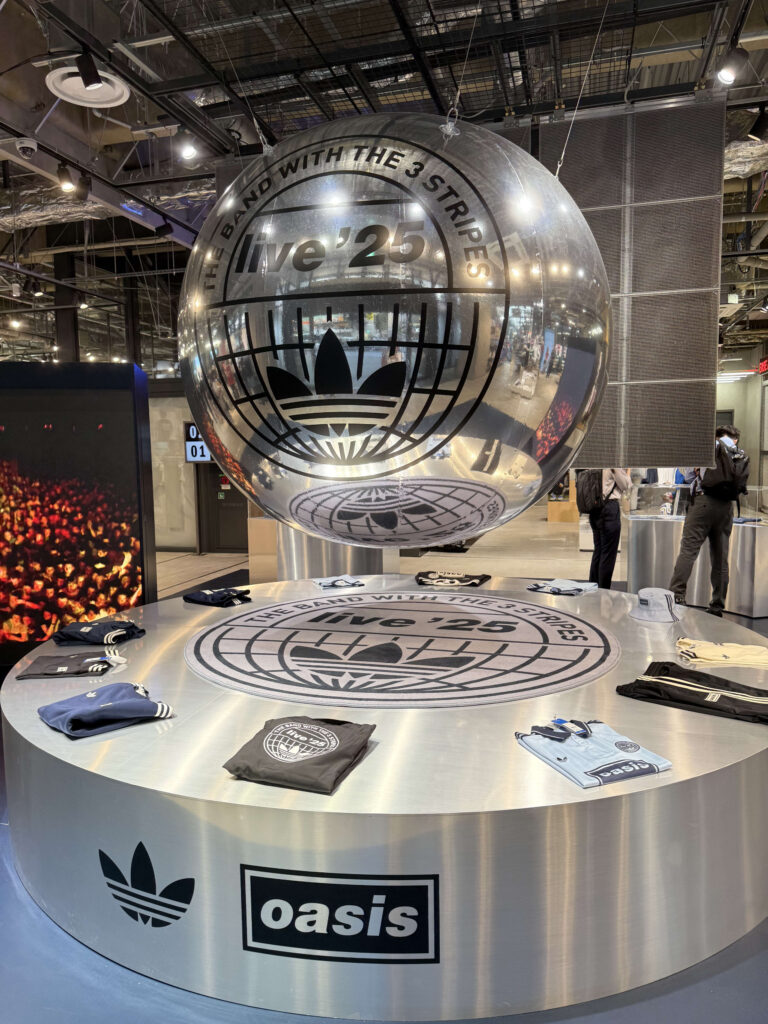 An in-store display for the Adidas and Oasis 'Live '25' collaboration, featuring a large, silver sphere with a logo hanging in the center above a circular table showcasing collaboration t-shirts.