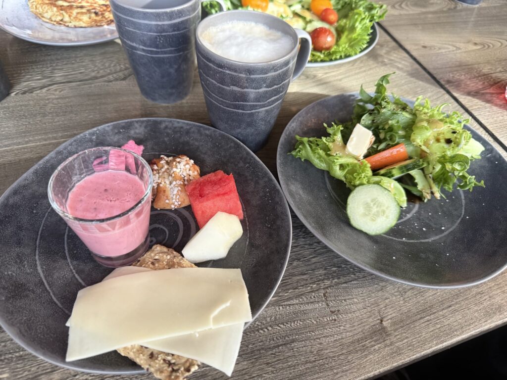 木製のテーブルの上に並べられた、彩り豊かな朝食。プレートにはピンクのスムージー、シナモンロール、フルーツ、チーズとクラッカー、サラダが盛り付けられ、横には泡立てられたミルクが入ったコーヒーが置かれている。