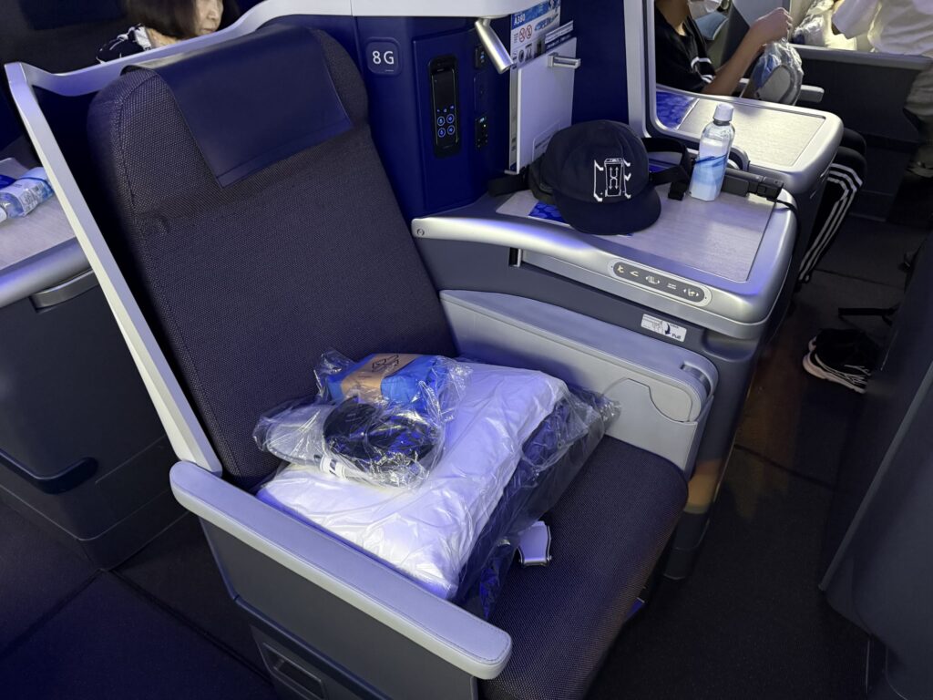 飛行機のビジネスクラスの座席。シートには枕やアメニティが置かれ、サイドテーブルには帽子と水が置かれている。