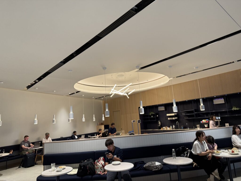 スタイリッシュな空港ラウンジの内部。多くの利用者が紺色のソファに座り、ノートパソコンで作業したり、食事をしたりしてリラックスしている。