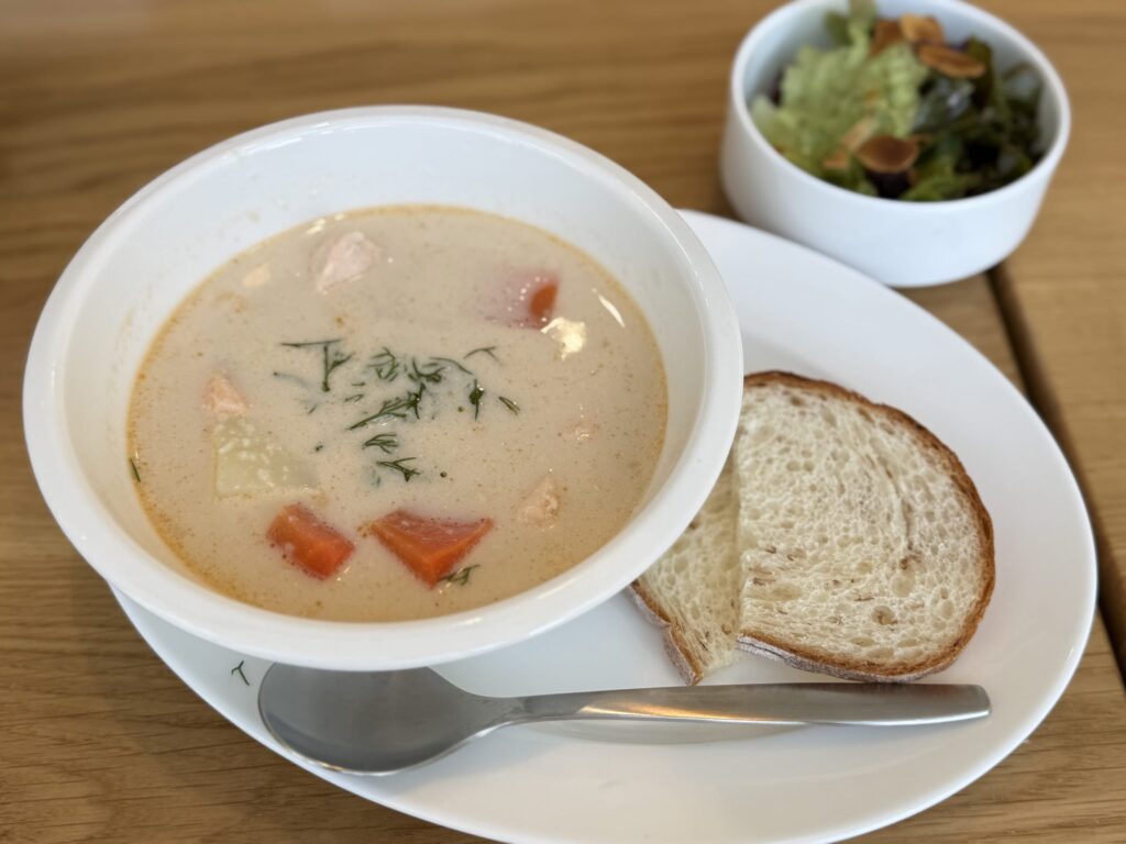 A lunch set on a wooden table, featuring a bowl of creamy salmon soup, bread, and a side salad. On a white plate sits a bowl of soup with hearty ingredients, sliced bread, and a salad topped with nuts.