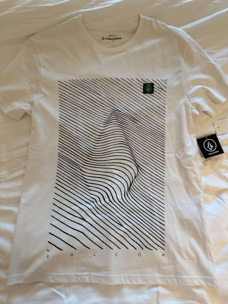 A white VOLCOM t-shirt laid flat, featuring a graphic of black parallel lines that are distorted to create a 3D optical illusion of a face.