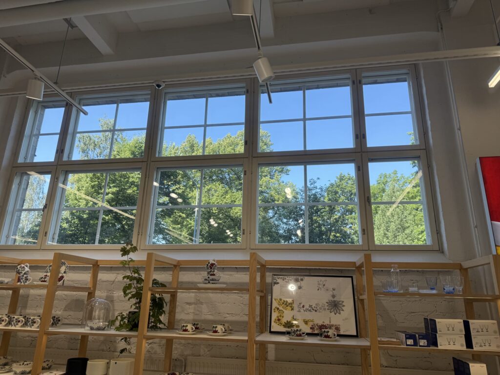 Large windows in a store look out onto green trees and a clear blue sky. In the foreground, wooden shelves are filled with ceramic tableware and glassware.