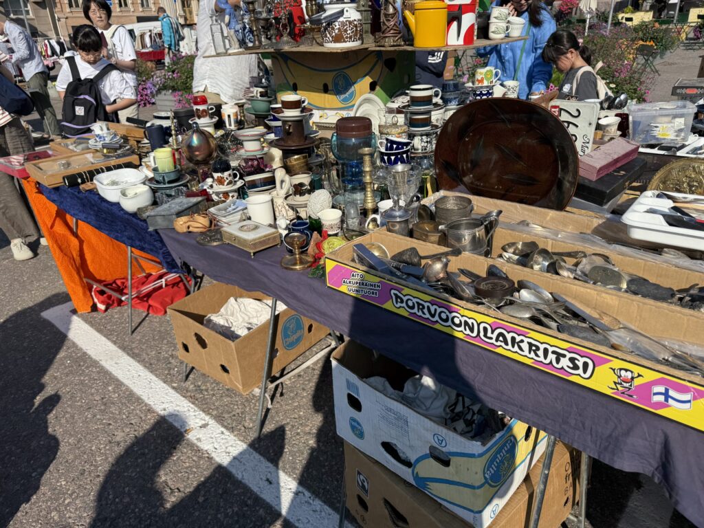 蚤の市で、ヴィンテージの食器やカトラリーがテーブルにずらりと並べられている。背景には買い物客の姿も見える。