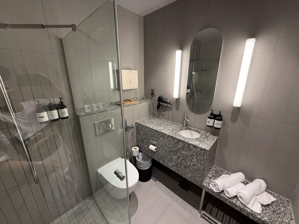 A modern hotel bathroom with grey tiles, featuring a glass-enclosed shower stall, a wall-mounted toilet, and a granite vanity.