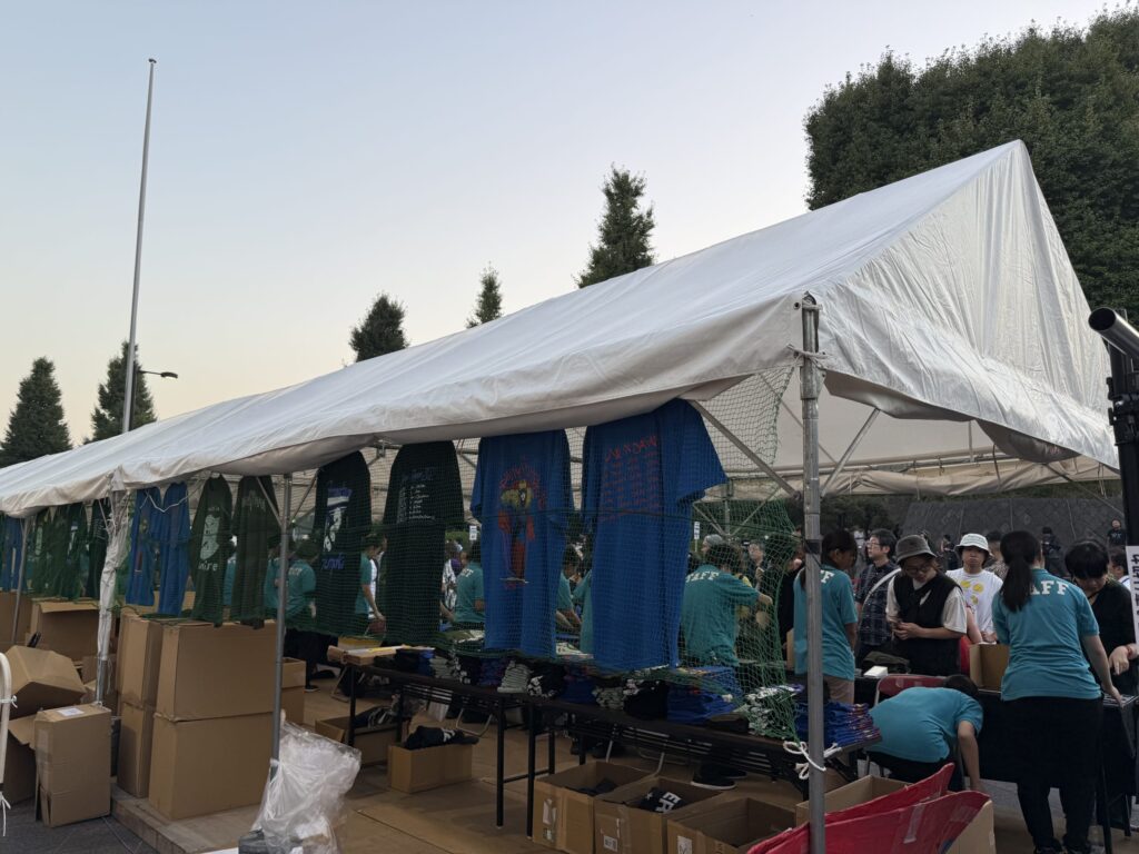 夕暮れ時の屋外イベント会場にあるグッズ販売のテント。陳列されたTシャツを見るために人々が集まっている。