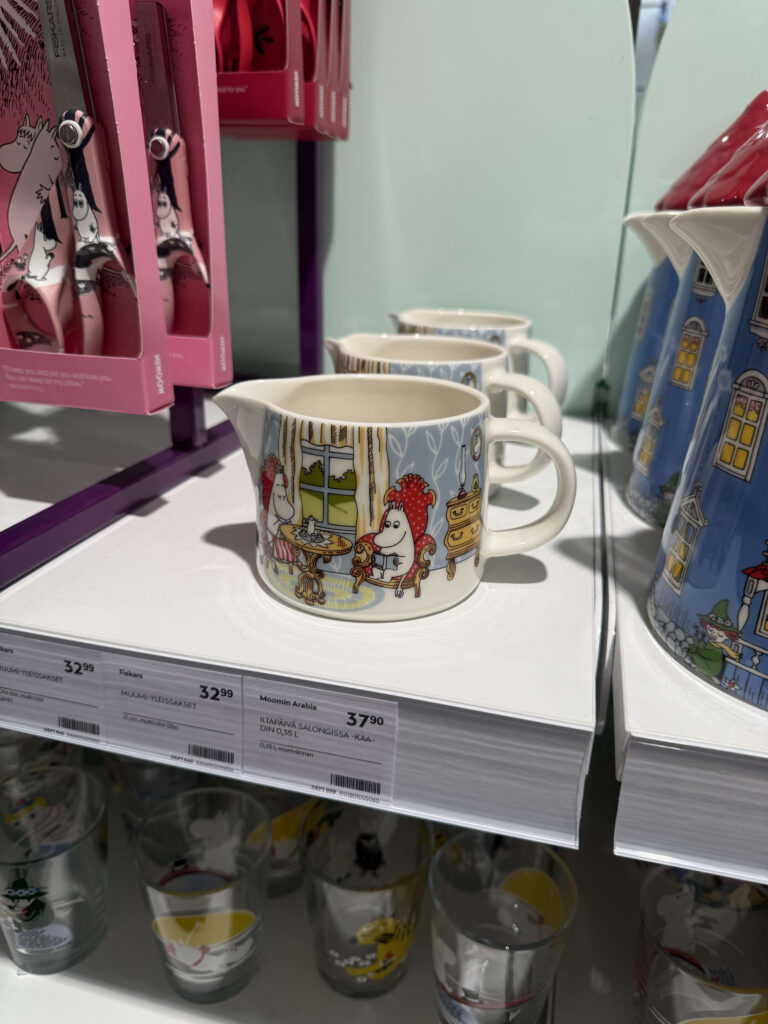 Moomin merchandise displayed on a department store shelf. In the foreground is a pitcher with an illustration of Moomintroll and Moominmamma, with Moominhouse-shaped mugs and Moomin-themed scissors in the background.