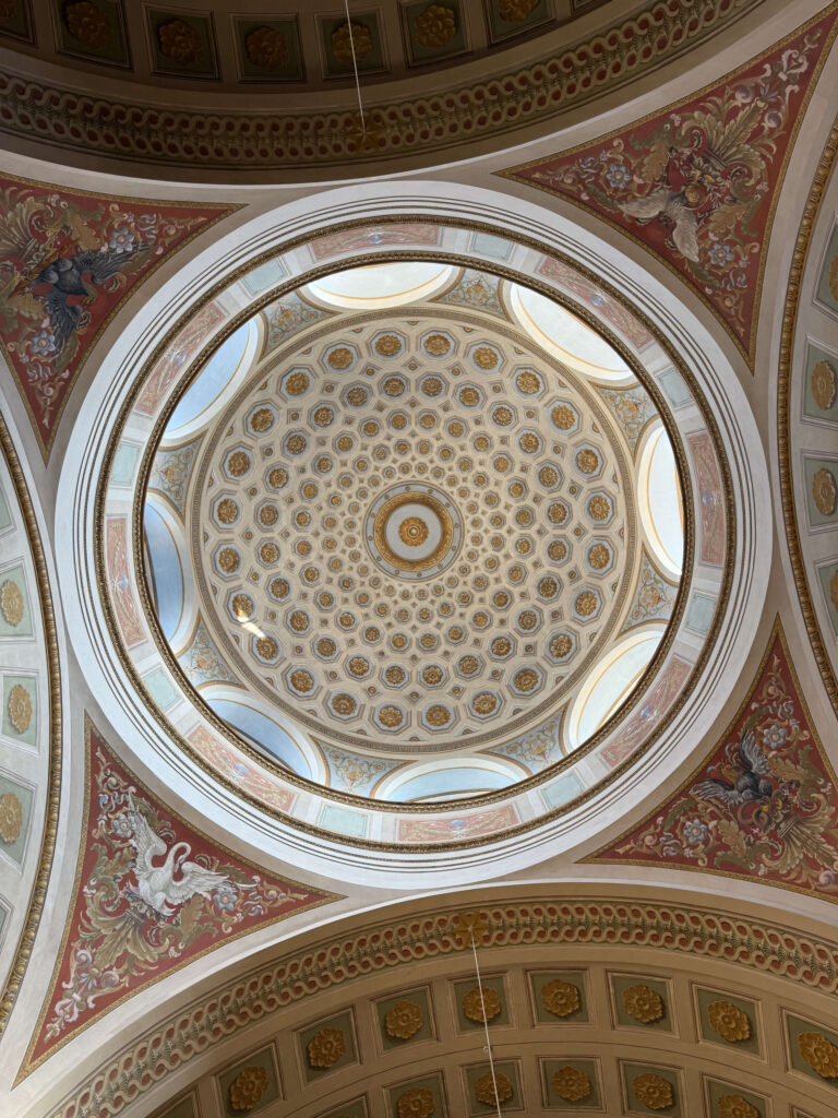 A photo looking directly up at an ornate, domed ceiling. The center features a coffered ceiling with golden floral decorations, and the surrounding triangular spaces are adorned with paintings of swans and eagles.