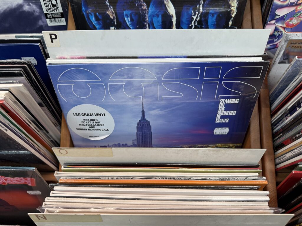 A vinyl record of the Oasis album 'Standing on the Shoulder of Giants' displayed in a record store bin. The cover features the New York City skyline at dusk.