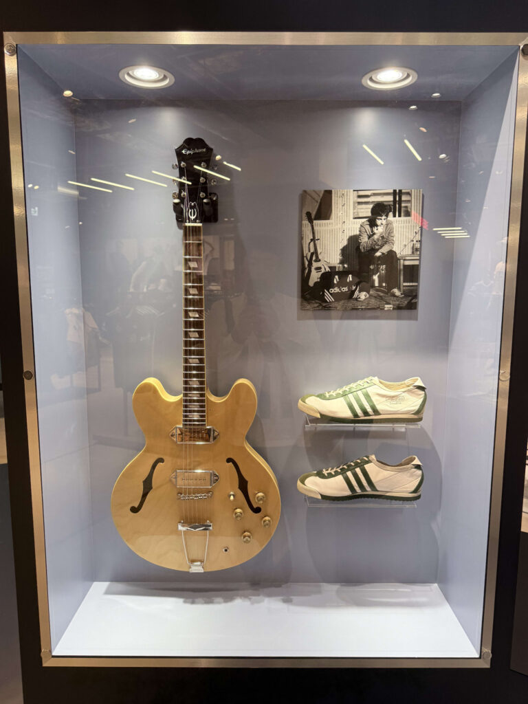A natural finish Epiphone electric guitar, a pair of white and green Adidas sneakers, and a black and white photograph of a musician displayed inside a glass case.