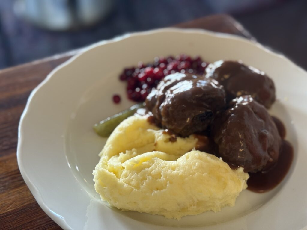 A white plate of Swedish meatballs covered in gravy, with mashed potatoes, lingonberry jam, and a pickle on the side.