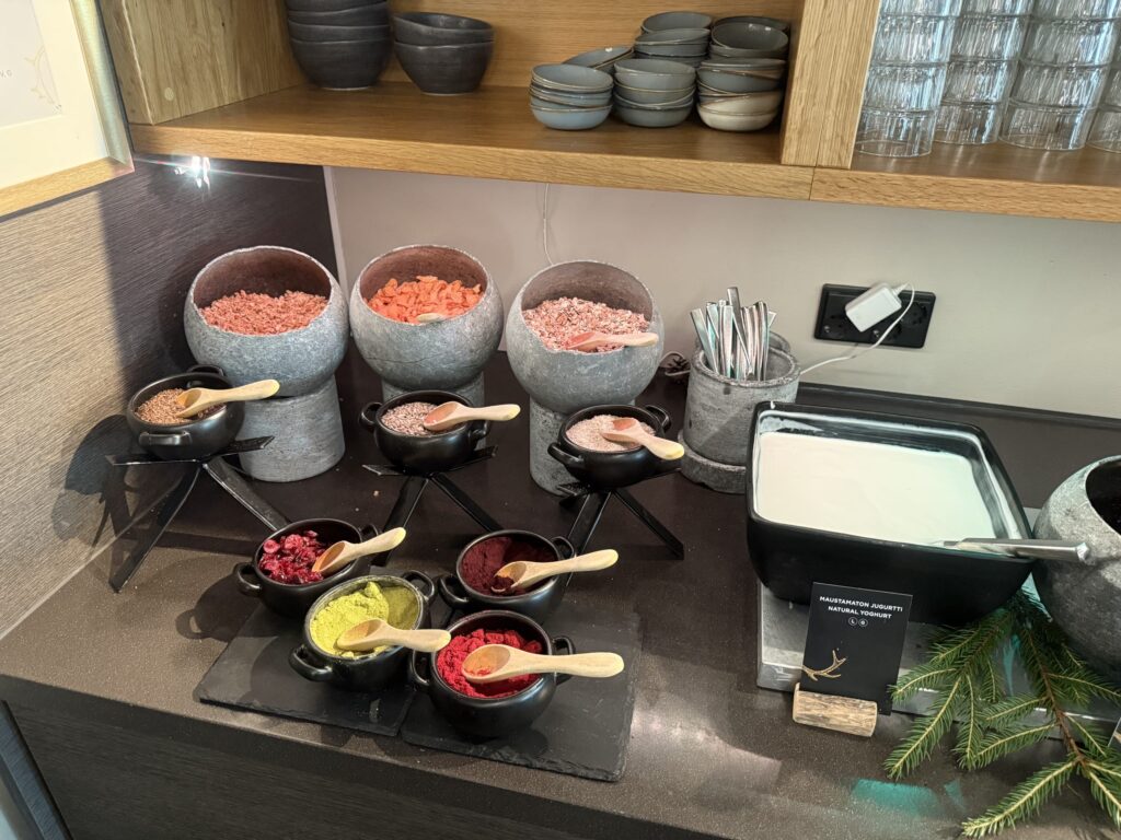 A yogurt bar at a hotel buffet. A wide variety of toppings such as cereals, seeds, and colorful powders are arranged in stone and black bowls.