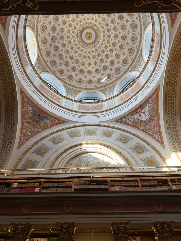 An upward shot of a magnificent dome ceiling in a library. It is decorated with golden geometric patterns and paintings of mythological creatures, with bookshelves visible at the bottom.