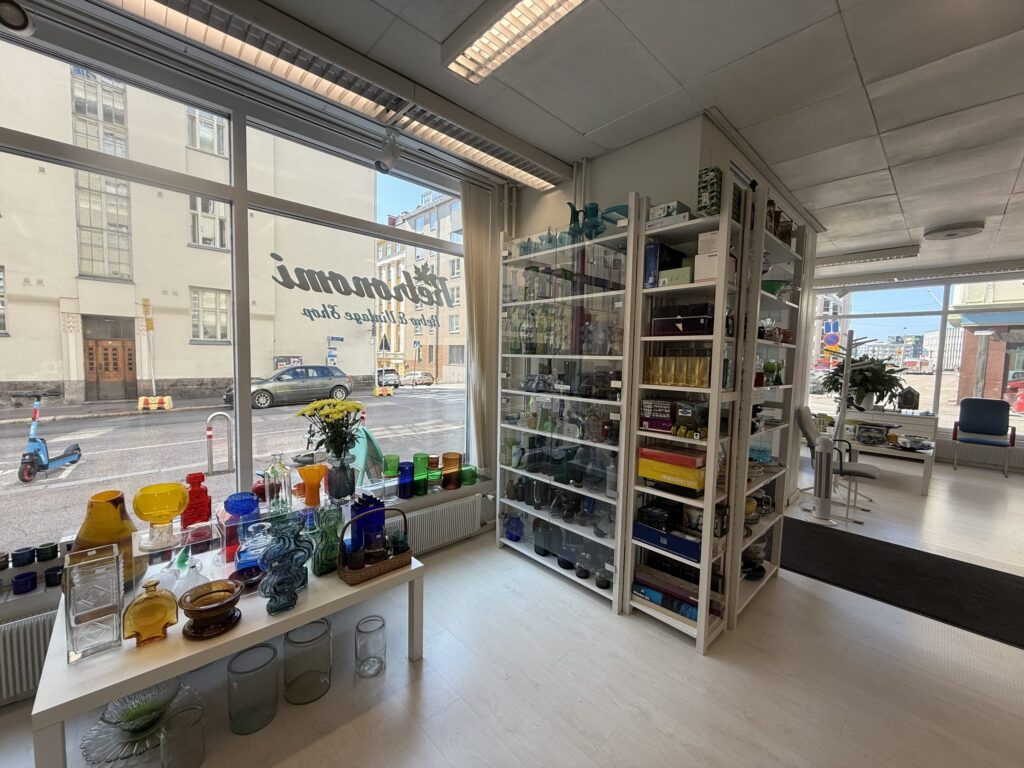 The interior of a sunlit vintage glassware shop. Colorful glass products are lined up on white shelves and tables by a large window overlooking a street.