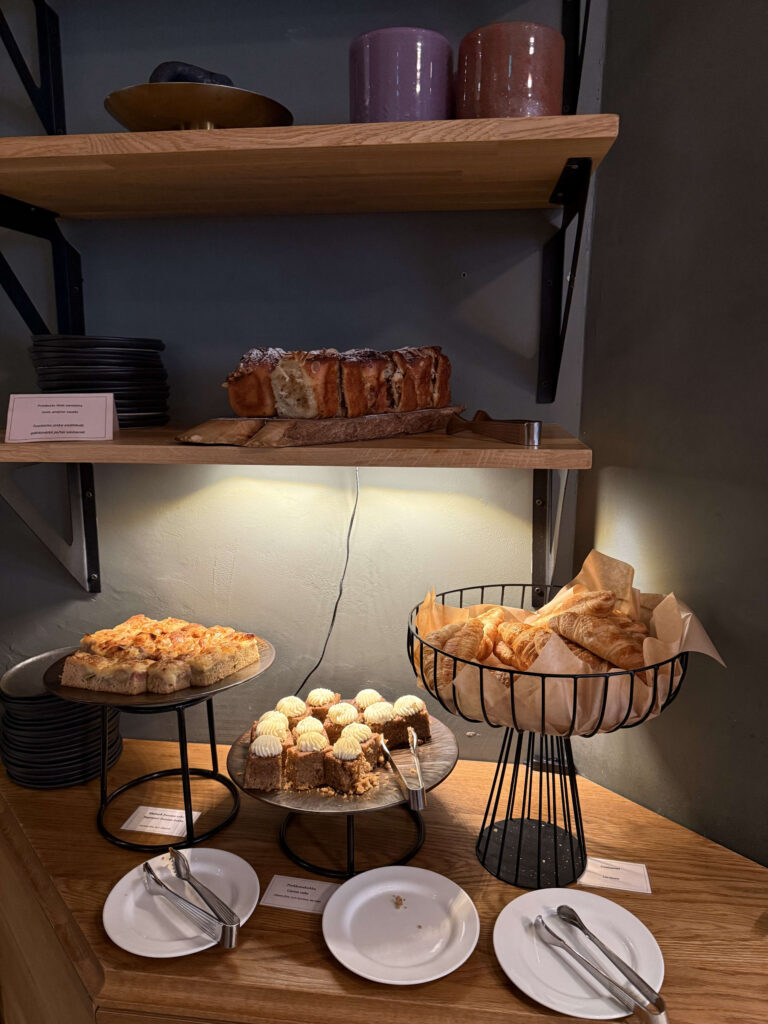 カフェの木製カウンターと棚に並べられた美味しそうなケーキ、パン、クロワッサン。ビュッフェ形式で、白いお皿とトングが用意されている。