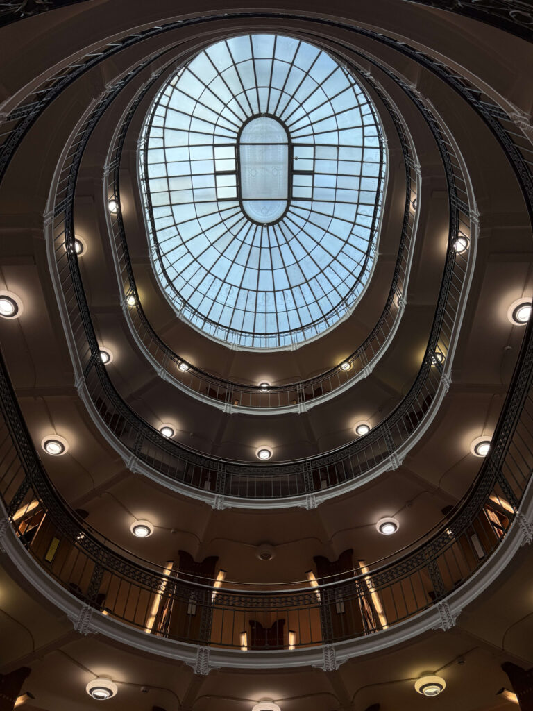 A low-angle shot looking up through a multi-story oval atrium. At the top is a large glass skylight, and the corridors of each floor feature decorative railings and warm-colored lighting.