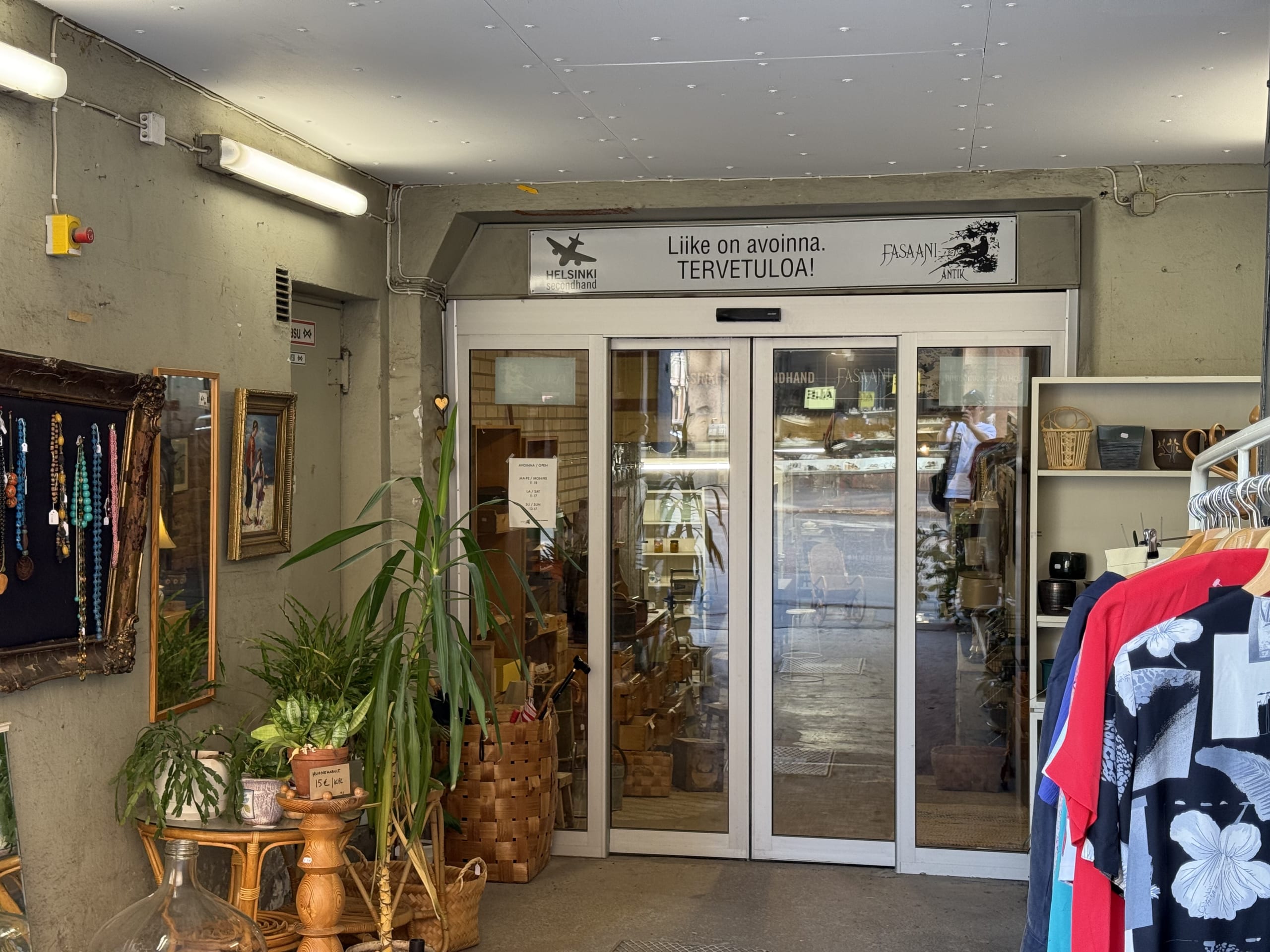 The entrance to a second-hand shop in Helsinki, with plants, antique goods, and clothing displayed around the automatic glass doors.