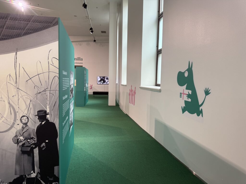 View of a Moomin exhibition hall with a green floor. There are display panels on the left and a white wall with a Moomin character on the right.