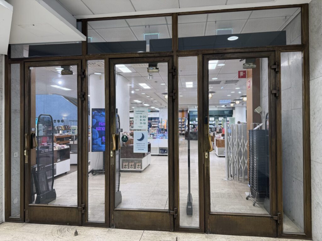 The side entrance of the Academic Bookstore, featuring four glass doors with wire mesh and heavy bronze-colored frames, through which the bright interior of the store is visible.