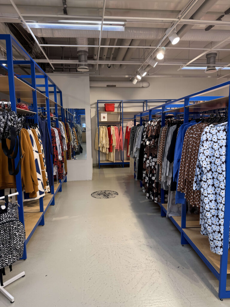 青いラックが両側に並ぶアパレルショップの通路。様々な色や柄の洋服が陳列されている。