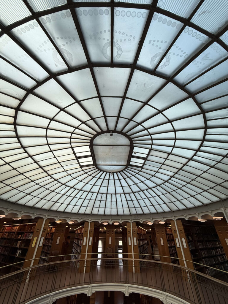 An upward-looking photo of a giant domed glass skylight with a grid-like iron frame, located in a circular library reading room. Bookshelves arranged in a circle are visible below.