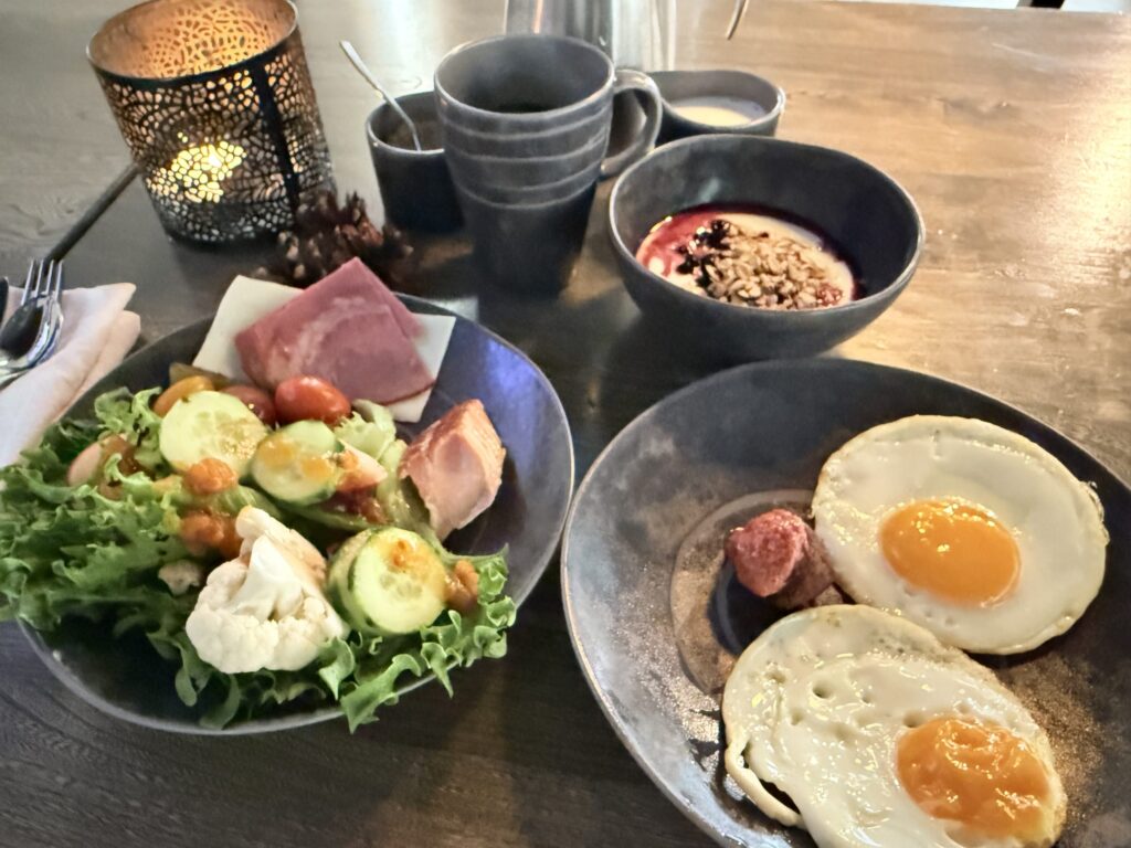 木製のテーブルの上に並べられた朝食。黒いお皿には、目玉焼きとソーセージ、レタスやきゅうりのサラダ、ハムとサーモン、そしてベリーソースとグラノーラがかかったヨーグルトが盛り付けられている。
