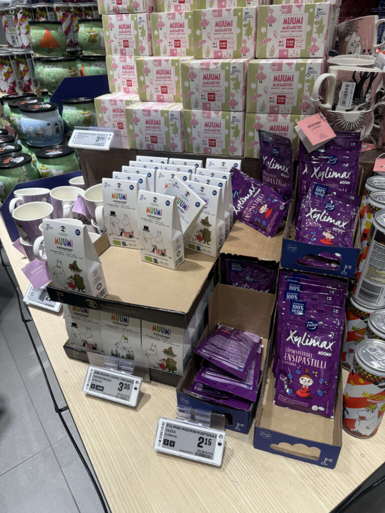 A display shelf in a supermarket showing various Moomin-designed products such as tea, chocolate, sweets, and mugs.