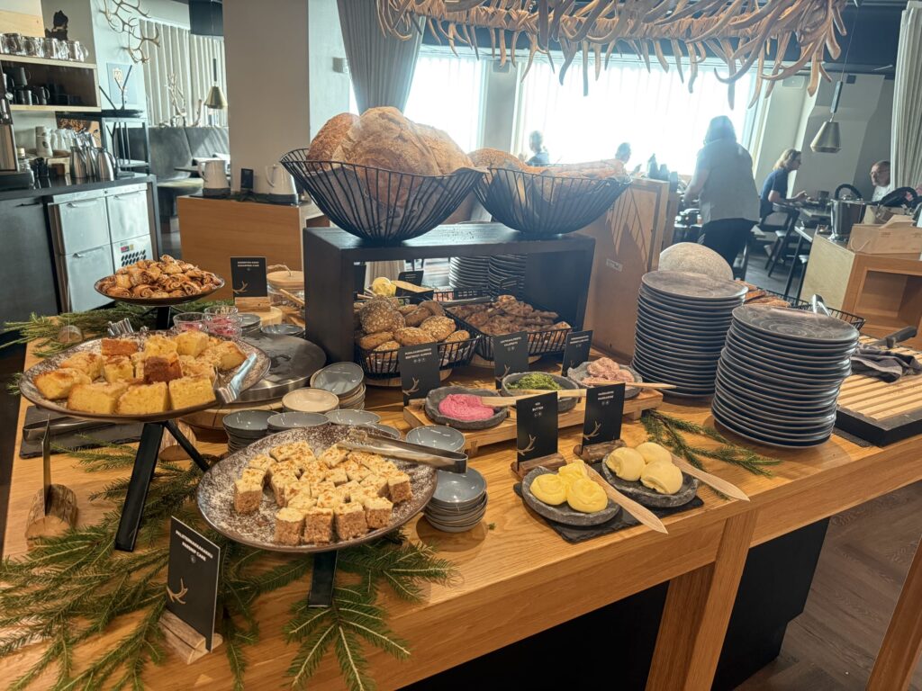 レストランの木製テーブルに並べられた朝食ビュッフェ。パン、ケーキ、スプレッド、バターなどがきれいに盛り付けられ、背景には鹿の角のシャンデリアが見える。