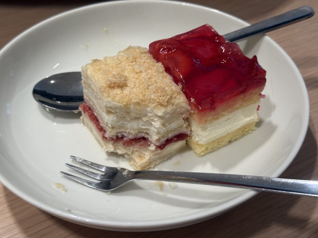 Two slices of cake, a mille-feuille and a red jelly-topped cake, are placed on a white plate with a fork and spoon.