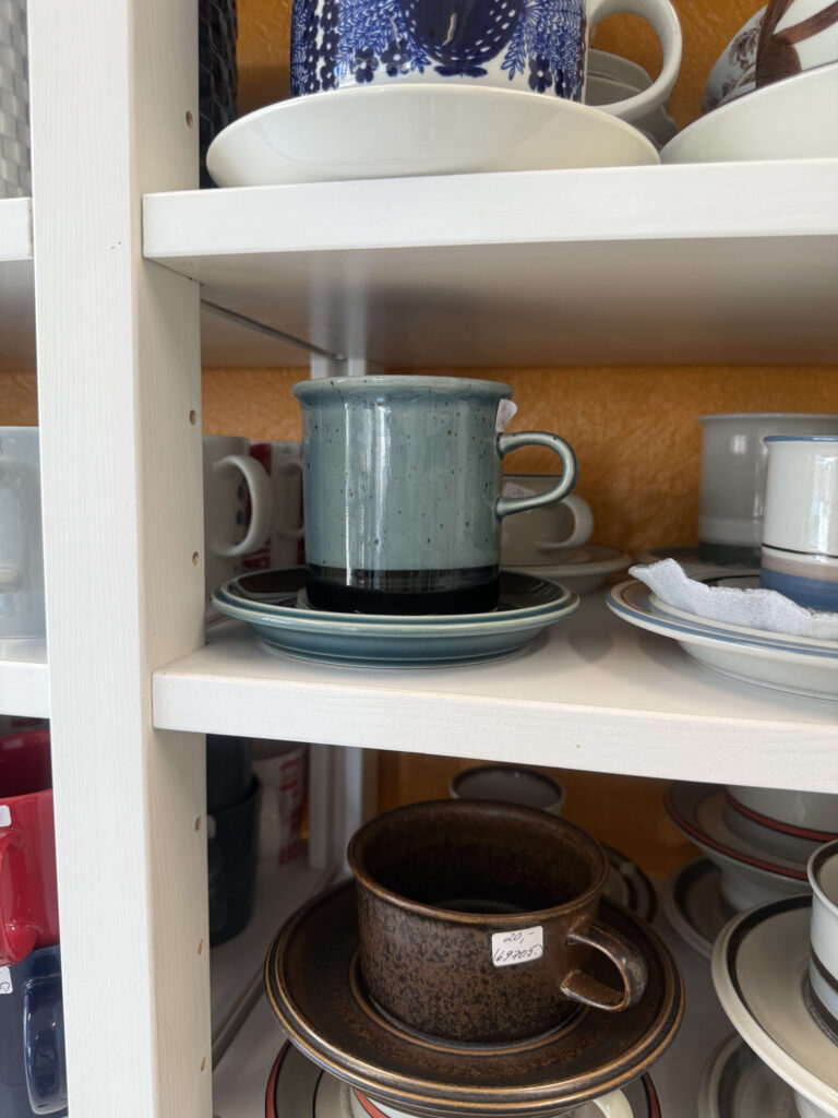 白い棚に置かれた、深みのある青緑色のコーヒーカップとソーサー。上下の棚にも、茶色や青い模様のヴィンテージ食器が並んでいる。
