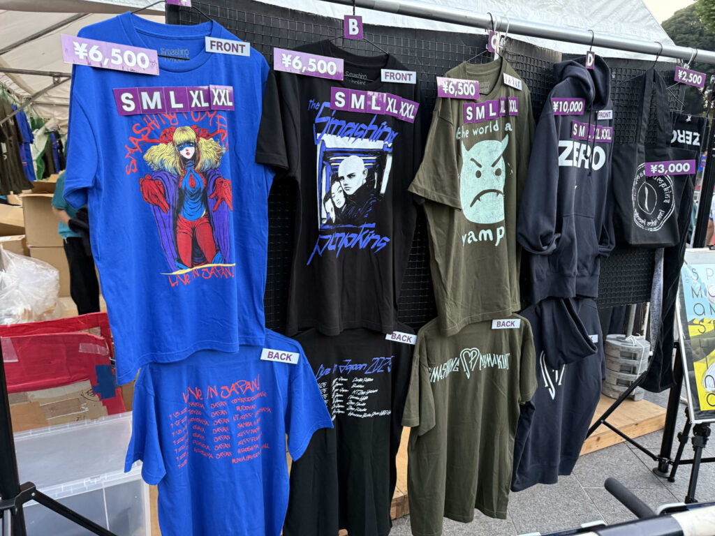 屋外の物販ブースに並べられたスマッシング・パンプキンズのコンサートグッズ。様々なデザインのTシャツやパーカーがハンガーにかけられており、値段とサイズの札が付いている。