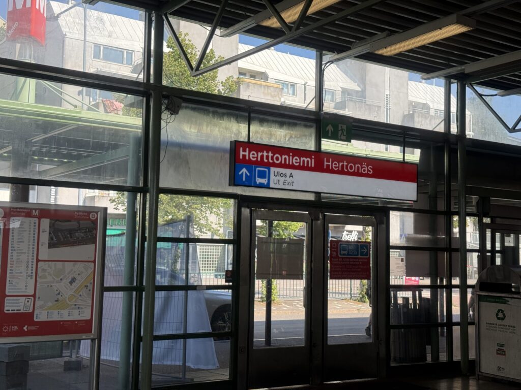 Inside Herttoniemi station in Helsinki. A red and white sign reading 'Herttoniemi Herttonäs' and indicating 'Ulos A / Ut Exit' hangs above glass doors.
