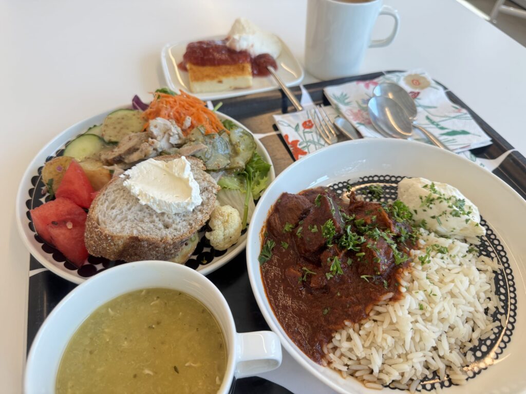 A complete meal on a tray: beef curry and rice, chicken and vegetable salad, bread, watermelon, soup, strawberry cheesecake, and coffee, served on Marimekko-style dishes.