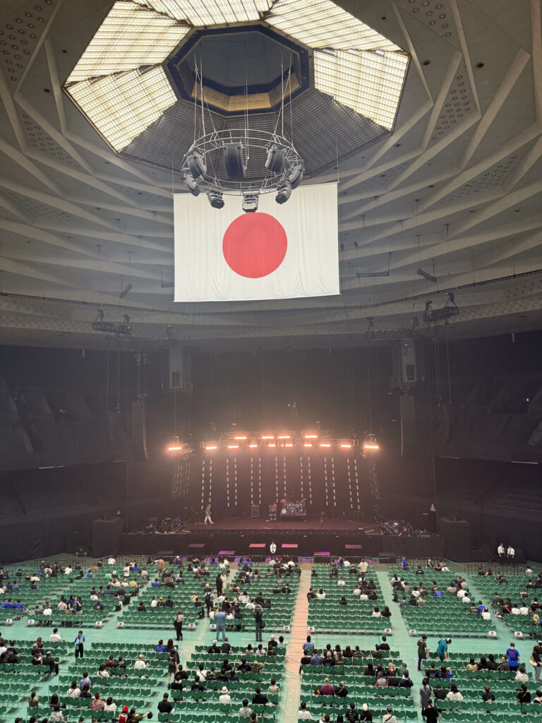 日本武道館の2階席からステージとアリーナ席を見下ろした俯瞰写真。八角形の天井から大きな日の丸が吊るされ、ステージには照明と楽器がセットされている。緑色の椅子が並んだアリーナ席には、開演を待つ観客が座り始めている。