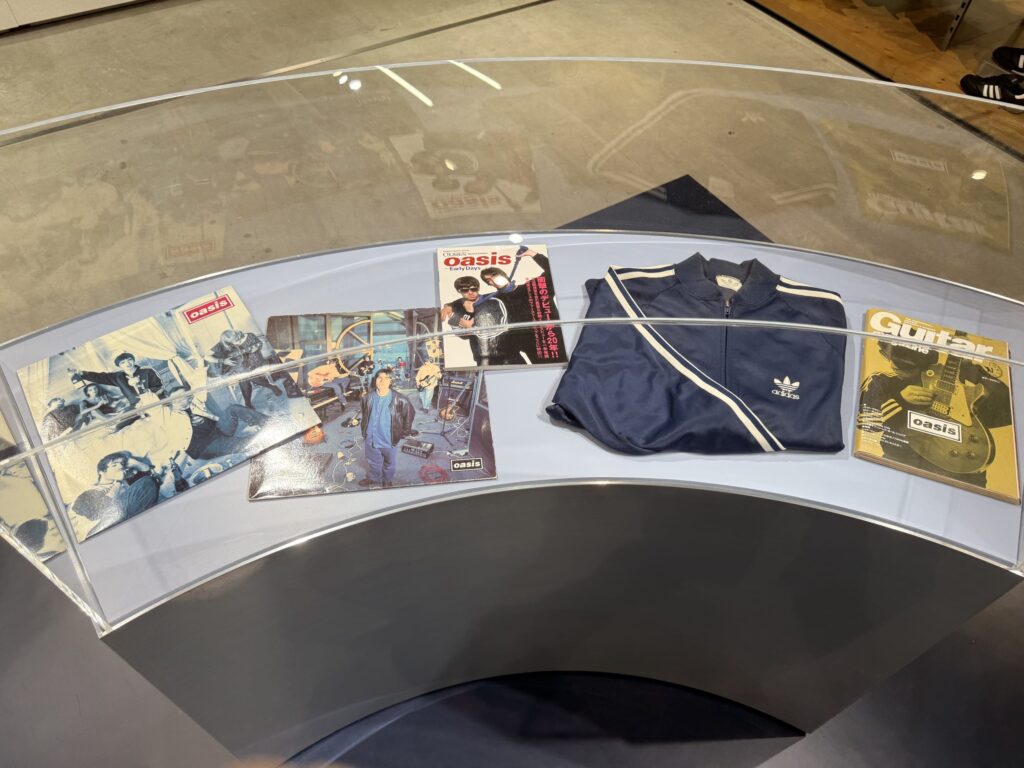 Memorabilia of the British rock band Oasis displayed inside a curved glass case, including a navy blue Adidas track jacket, record sleeves, and music magazines.