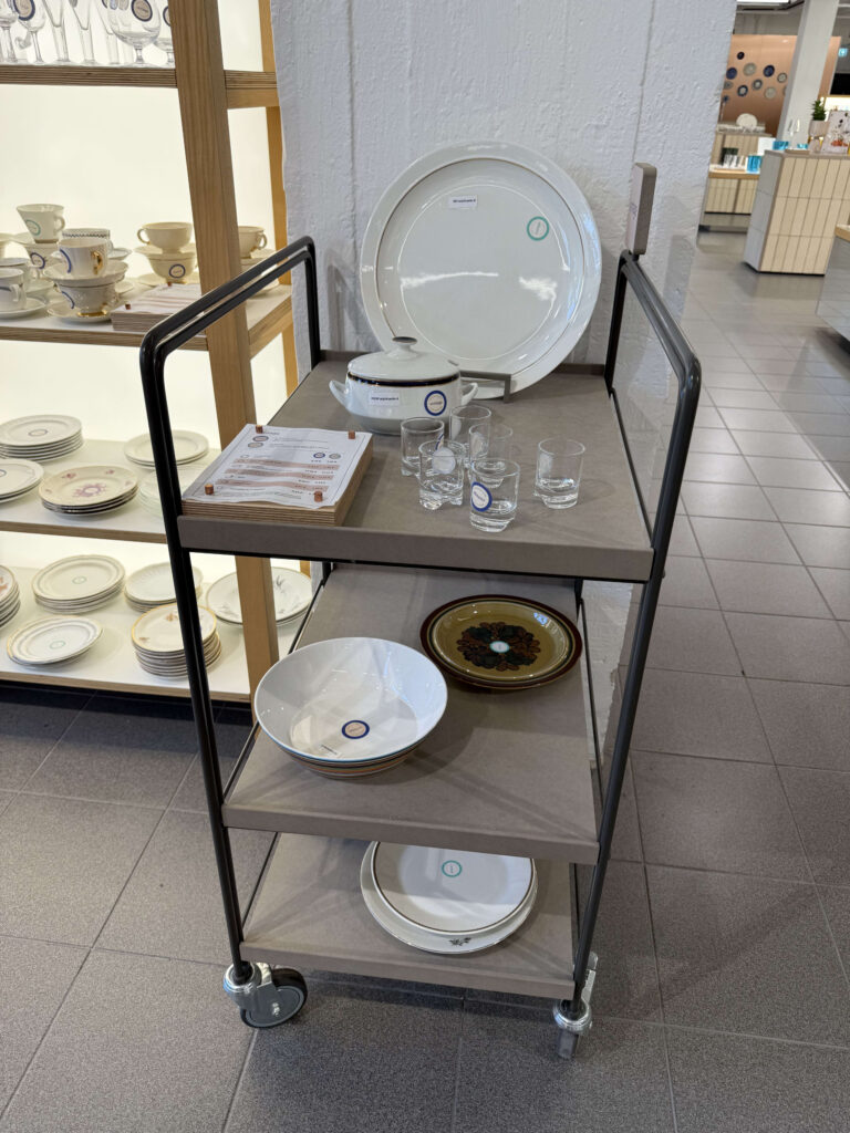 A three-tiered cart with a black frame in a tableware store. The top shelf holds a large white platter, a lidded tureen, and glasses, while the middle and bottom shelves display various bowls and plates.