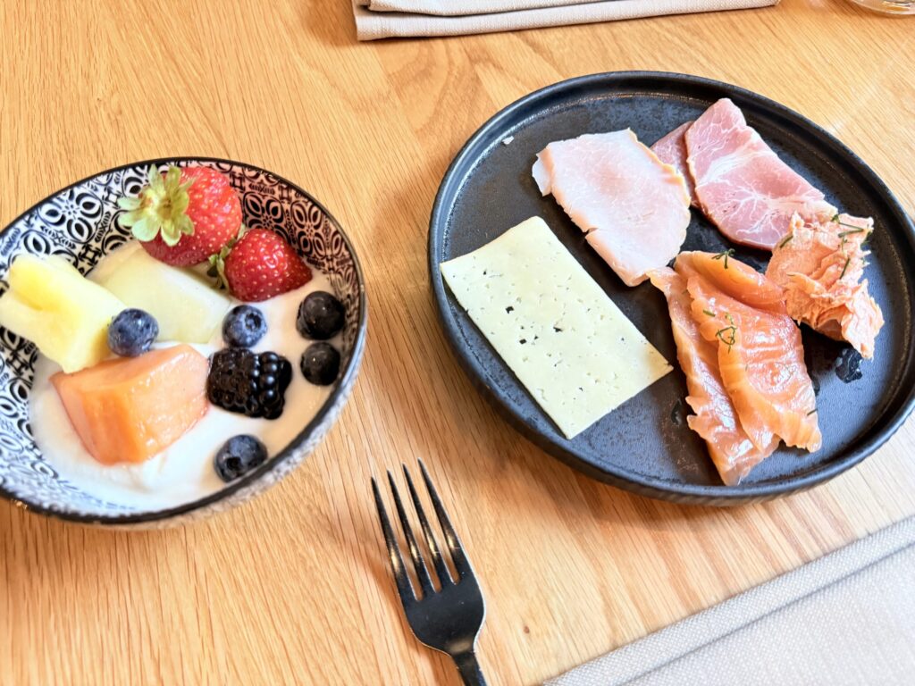 木製のテーブルの上に、フルーツとヨーグルトが入ったボウルと、ハム、チーズ、スモークサーモンが盛り付けられた黒い皿が置かれている。