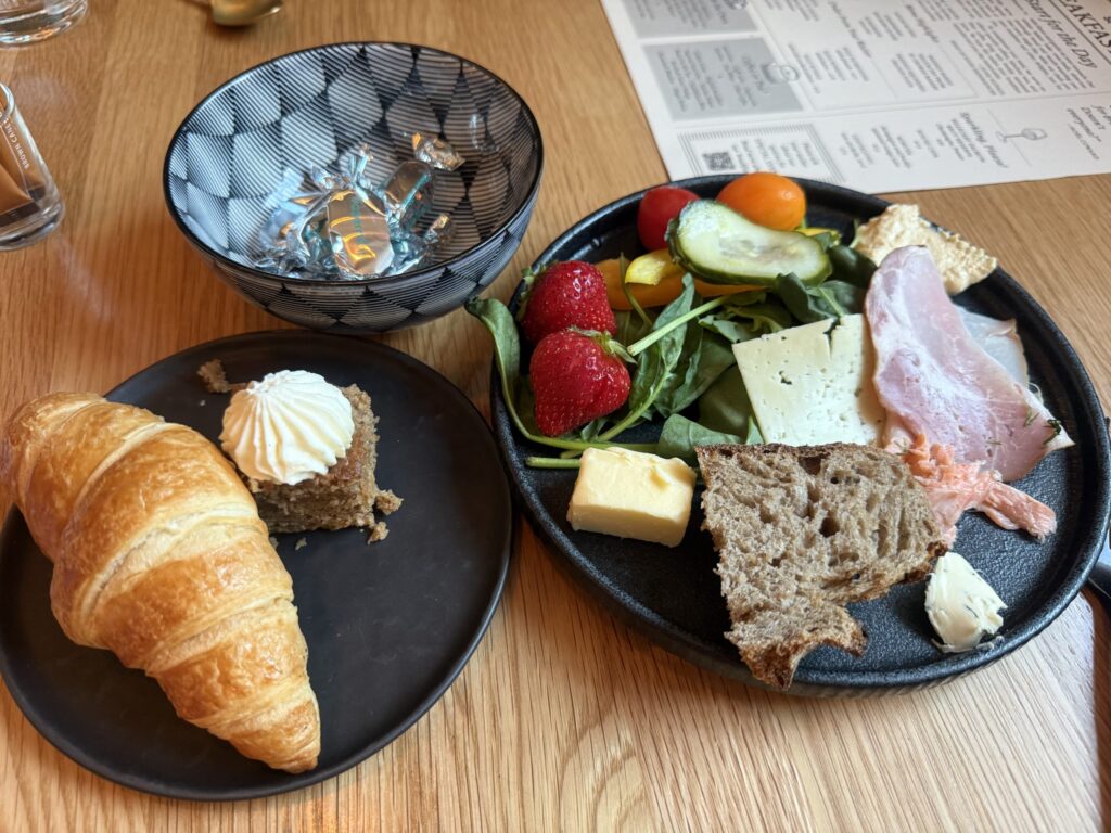 Two black plates on a wooden table. One plate holds ham, cheese, smoked salmon, a salad with strawberries and vegetables, and rye bread. The other plate has a croissant and a small cake. In the background is a geometric-patterned bowl with candies.